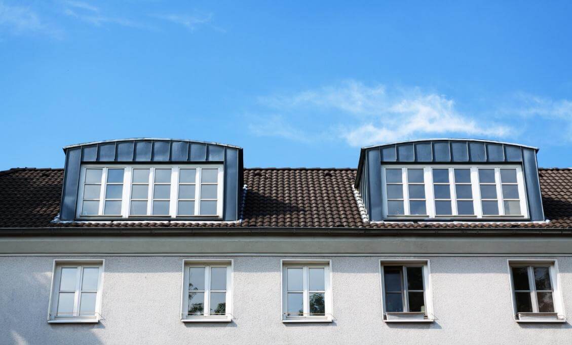 Dachgeschoss mit zwei Gauben und weißen Sprossenfenstern auf einem Wohnhaus mit Ziegeldach unter blauem Himmel