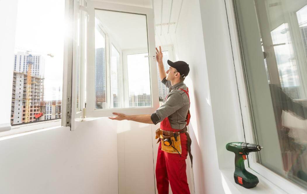 Handwerker in roter Arbeitskleidung montiert ein weißes Kunststofffenster in einer Wohnung, Akkuschrauber liegt auf der Fensterbank.