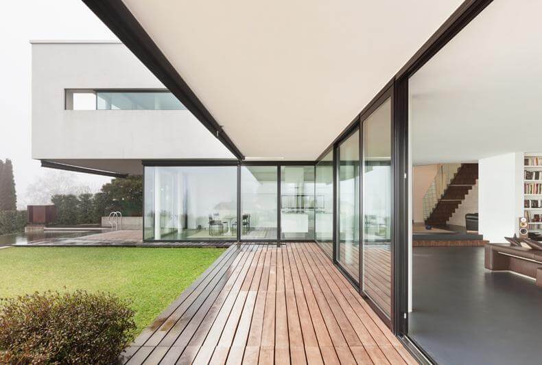 Moderne Villa mit bodentiefen Glasfronten, Holzterrasse und Blick in offenen Wohnbereich mit Treppe und Garten.