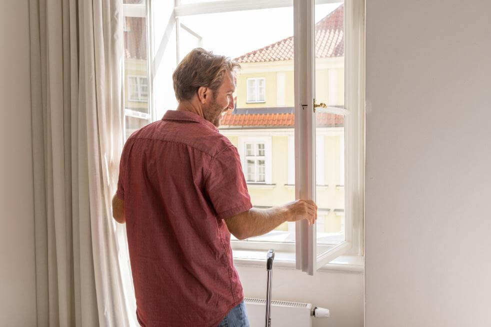 Mann öffnet ein Fenster in einem hellen Zimmer und blickt nach draußen