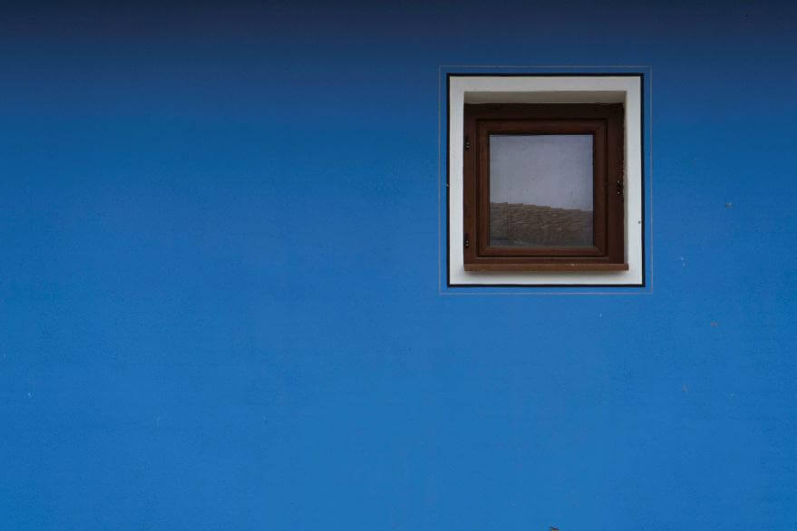 Kleines quadratisches Fenster mit braunem Rahmen in einer blauen Hauswand.