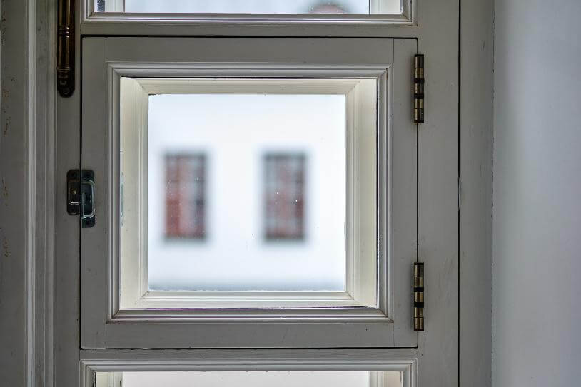 Fensterrahmen mit Griff und Scharnieren, Blick nach draußen auf eine unscharfe Fassade mit zwei Fenstern.
