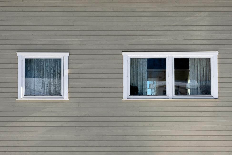 Hausfassade mit horizontaler Holzverkleidung und zwei weißen Fenstern mit Vorhängen.
