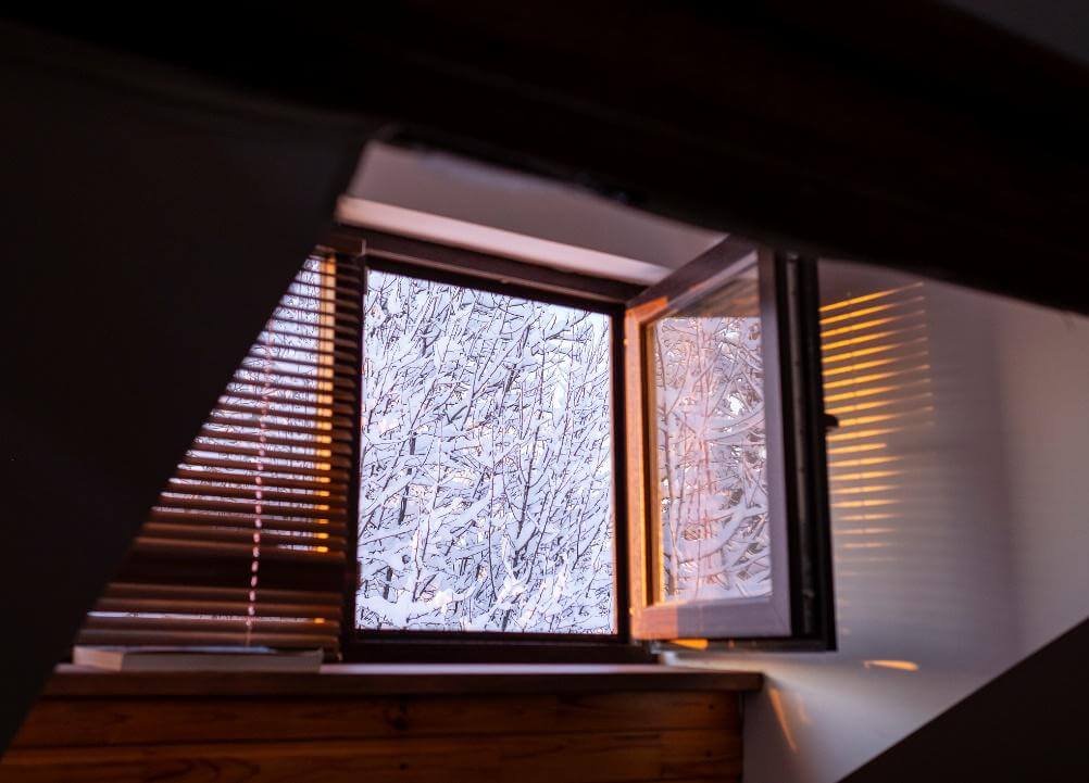 Geöffnetes Dachfenster mit Blick auf schneebedeckte Äste, innen Holzverkleidung und Jalousie.