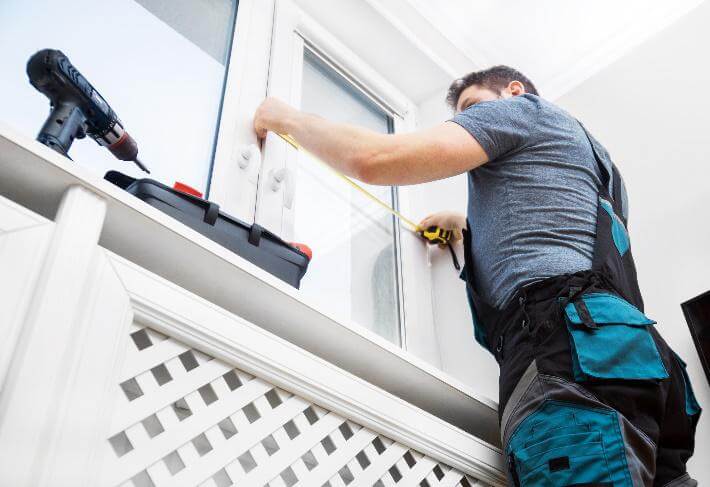 Monteur in Arbeitskleidung misst ein weißes Fenster im Innenbereich aus.