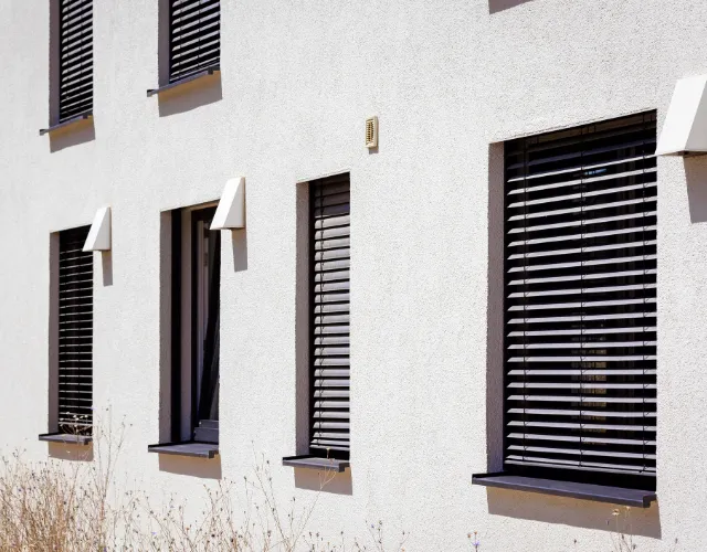 Dunkle Raffstores an der hellen Fassade eines Wohnhauses.