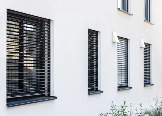 Schwarze Raffstores an mehreren Fenstern einer weißen Hausfassade