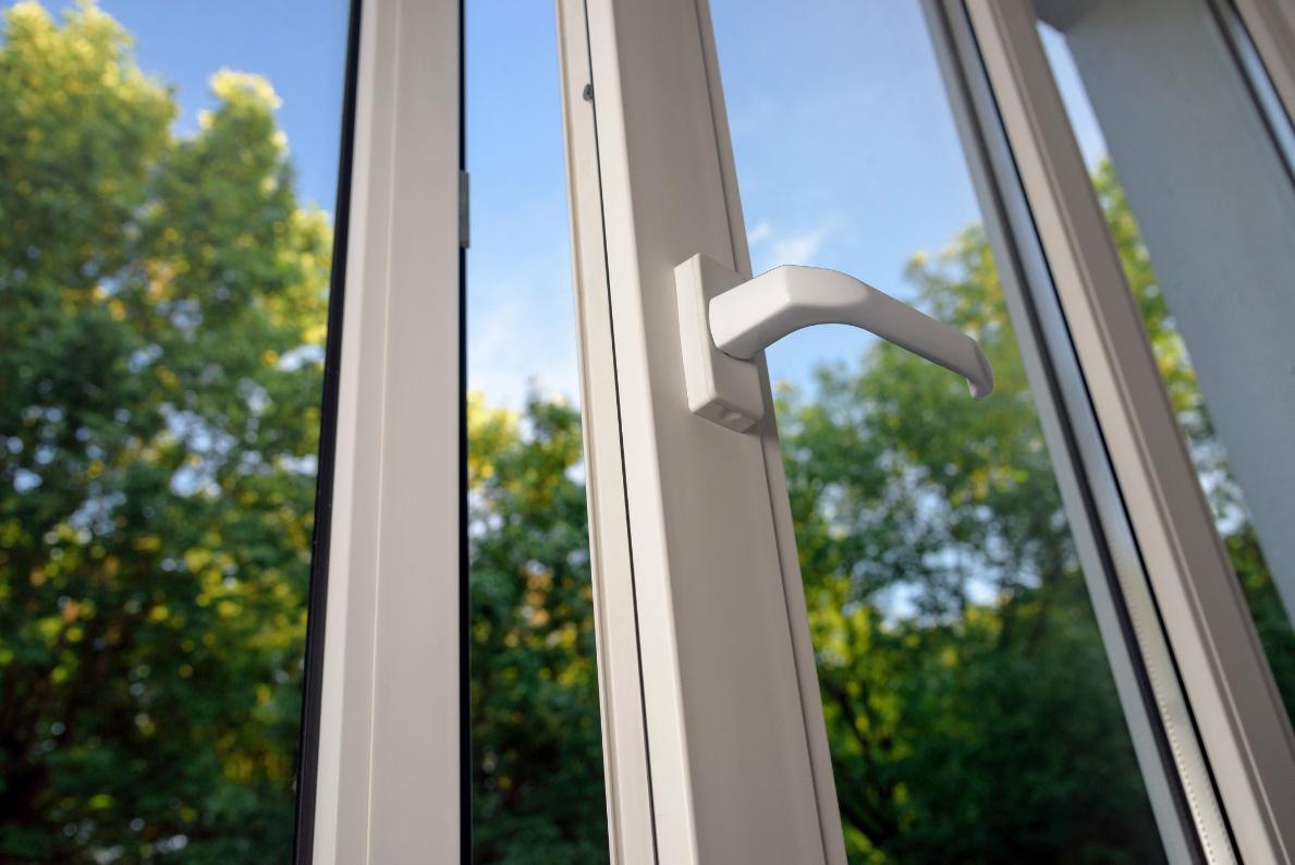Nahaufnahme eines geöffneten Fensters mit einem weißen Fenstergriff, durch das man auf grüne Bäume sieht.