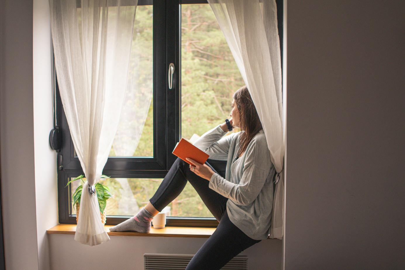 Eine Frau sitzt mit einem Buch in der Hand am Fenster und schaut hinaus.