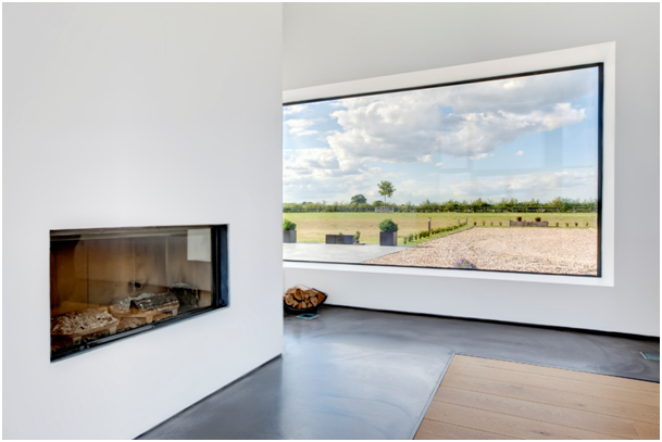 Heller Wohnraum mit integriertem Kamin, gro&szlig;em Panoramafenster und Blick auf eine weitl&auml;ufige Landschaft.