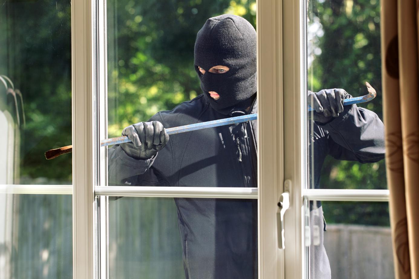 Person mit schwarzer Sturmhaube und Handschuhen hebelt mit einer Brechstange ein Fenster von au&szlig;en auf.