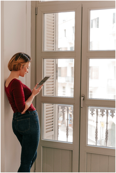 Junge Frau steht am geschlossenen Fenster und benutzt ein Tablet