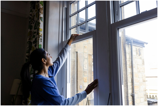 Eine Frau justiert den Fensterfl&uuml;gel eines Kunststofffensters. Der Ausblick f&auml;llt auf ein Geb&auml;ude.