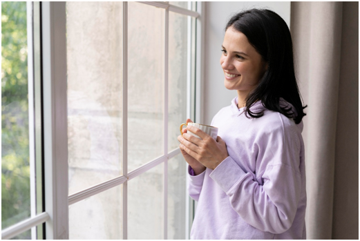 Frau h&auml;lt eine Tasse in der Hand und schaut gl&uuml;cklich aus dem geschlossenen Fenster