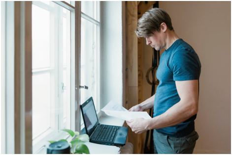Mann steht am Fenster und &uuml;berpr&uuml;ft Unterlagen vor einem Laptop