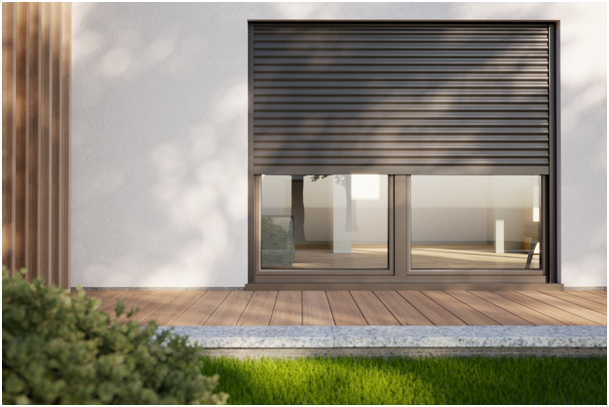 Teilweise geschlossener Rollladen &uuml;ber bodentiefem Fenster einer modernen Hausfassade mit Holzterrasse und Gartenbereich.