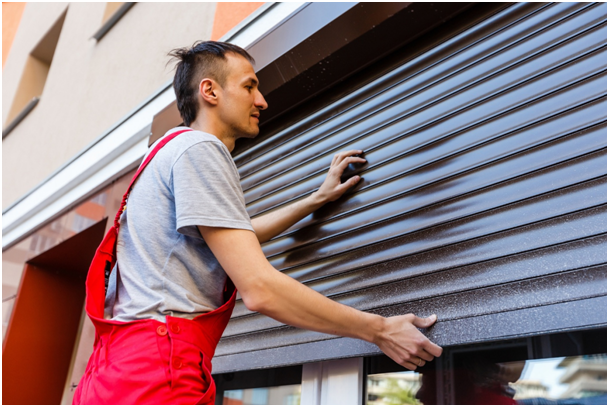 Handwerker in roter Arbeitskleidung richtet einen Rollladen an einer Hausfassade aus.