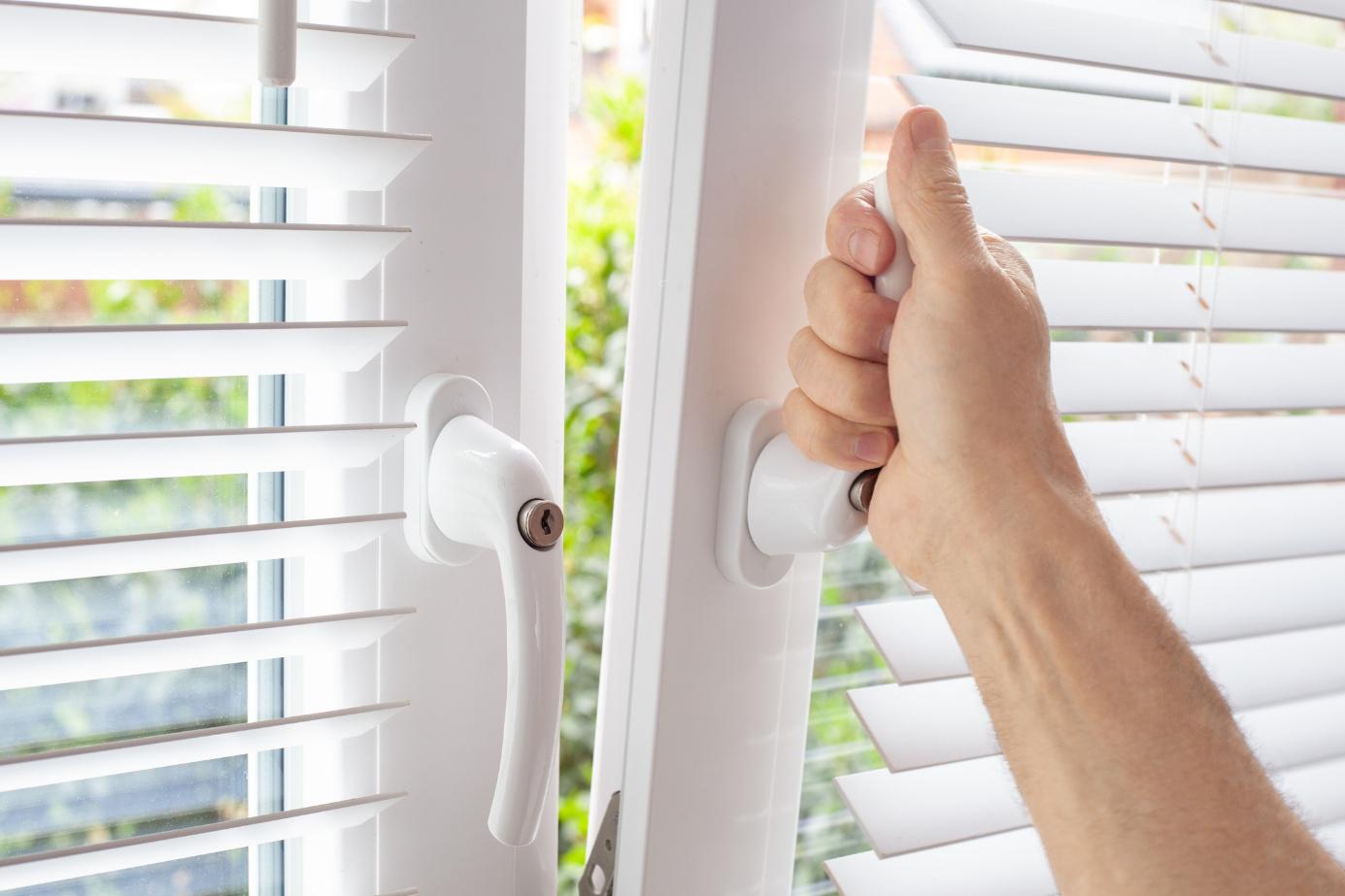 Nahaufnahme einer Hand, die einen Fenstergriff dreht, um das Fenster zu öffnen, mit weißen Jalousien im Hintergrund