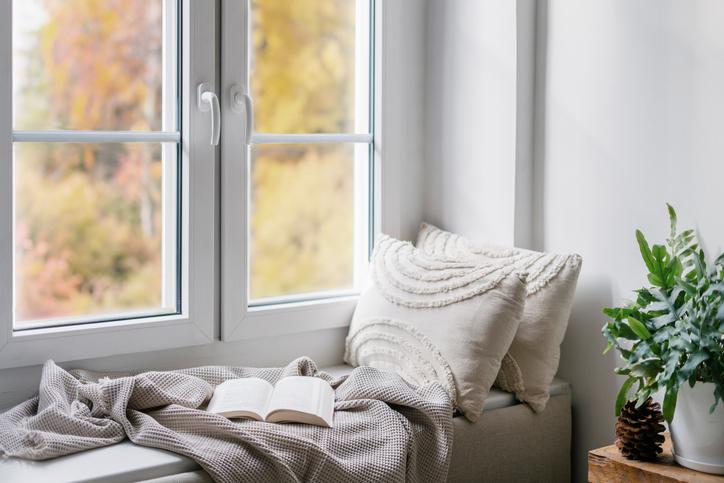 Fenster mit Blick auf Bäume, eine Sitzbank mit Kissen und Decke und geöffnetem Buch darauf.